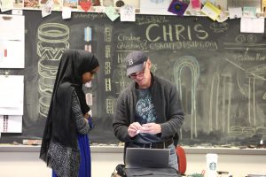 a student in a black head covering speaks to a teacher in a baseball cap in front of a chalkboard.