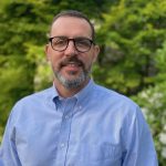 Tom Flanagan stands smiling, wearing a blue button-down shirt and glasses, in front of a tree