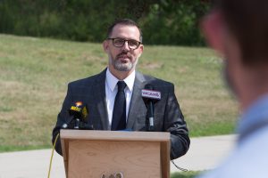 Tom Flanagan, wearing glasses and a suit, stands behind a podium during a press conference.